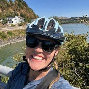 Smiling cyclist in helmet and sunglasses takes selfie by a river with a house and hills