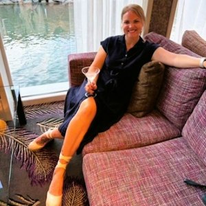 Smiling woman in navy dress reclining on purple sofa by window with waterfront view, holding martini