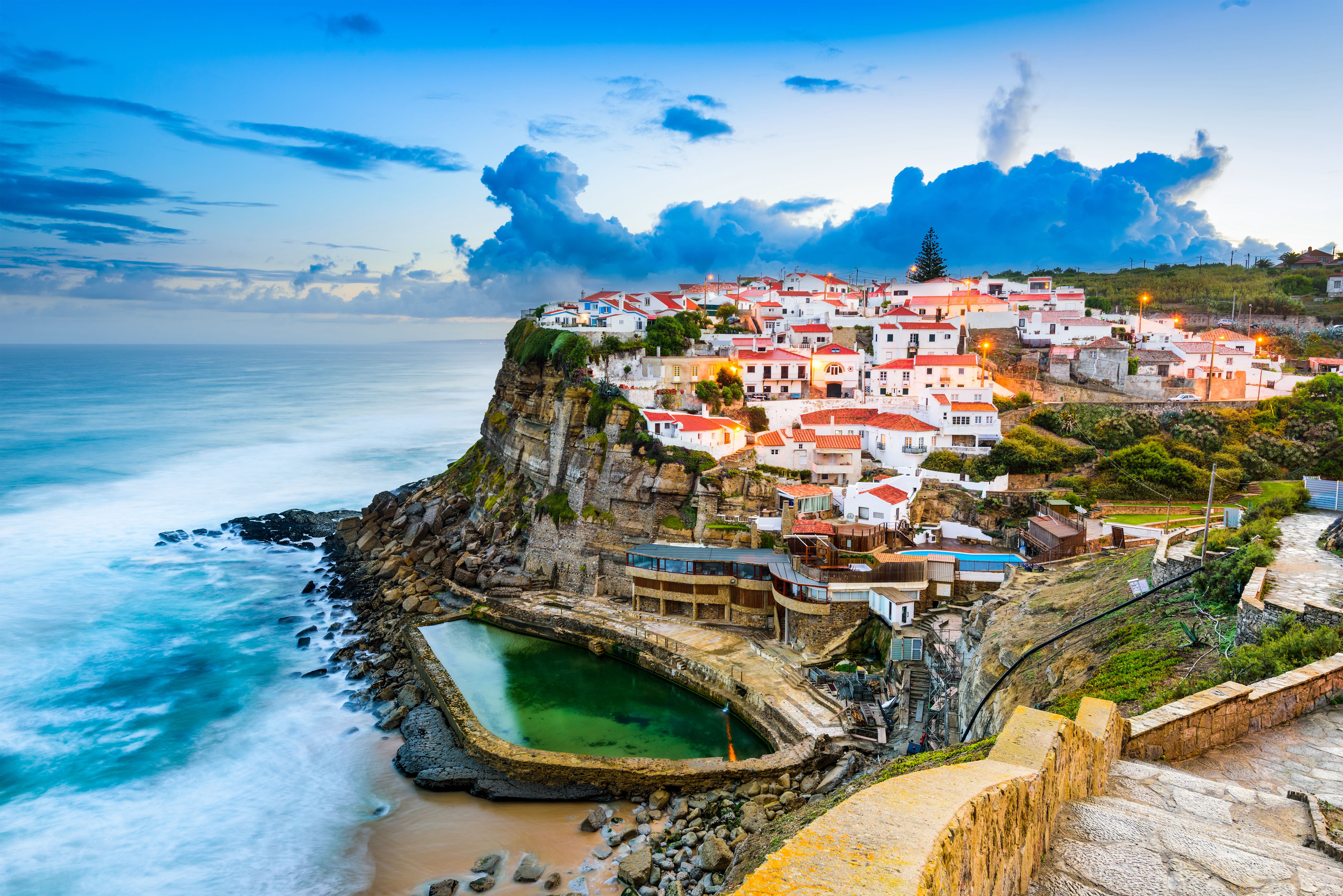 Clifftop village of white houses with red roofs, tidal pool and stone steps overlooking ocean.