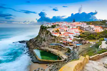Clifftop village of white houses with red roofs, tidal pool and stone steps overlooking ocean.