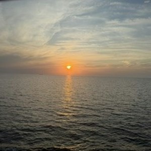 Sunset over open ocean, sun at horizon casting golden path on rippling water beneath wispy clouds