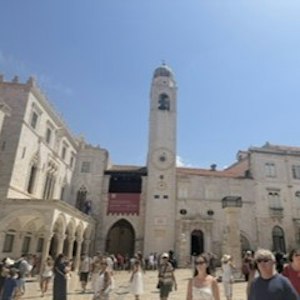 Sunlit historic stone plaza with tall bell tower, arched colonnades and tourists under blue sky