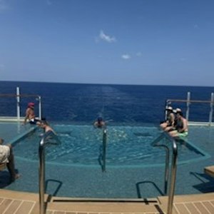 Infinity pool on cruise ship with swimmers at edge and glass rail overlooking open ocean horizon.