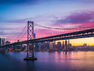Suspension bridge over bay with city skyline reflecting pink-purple sunset hues.