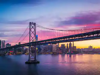 Suspension bridge over bay with city skyline reflecting pink-purple sunset hues.