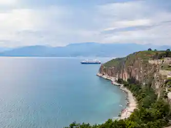 Cruise ship near cliffs and turquoise sea with tree-covered headland and distant mountains.