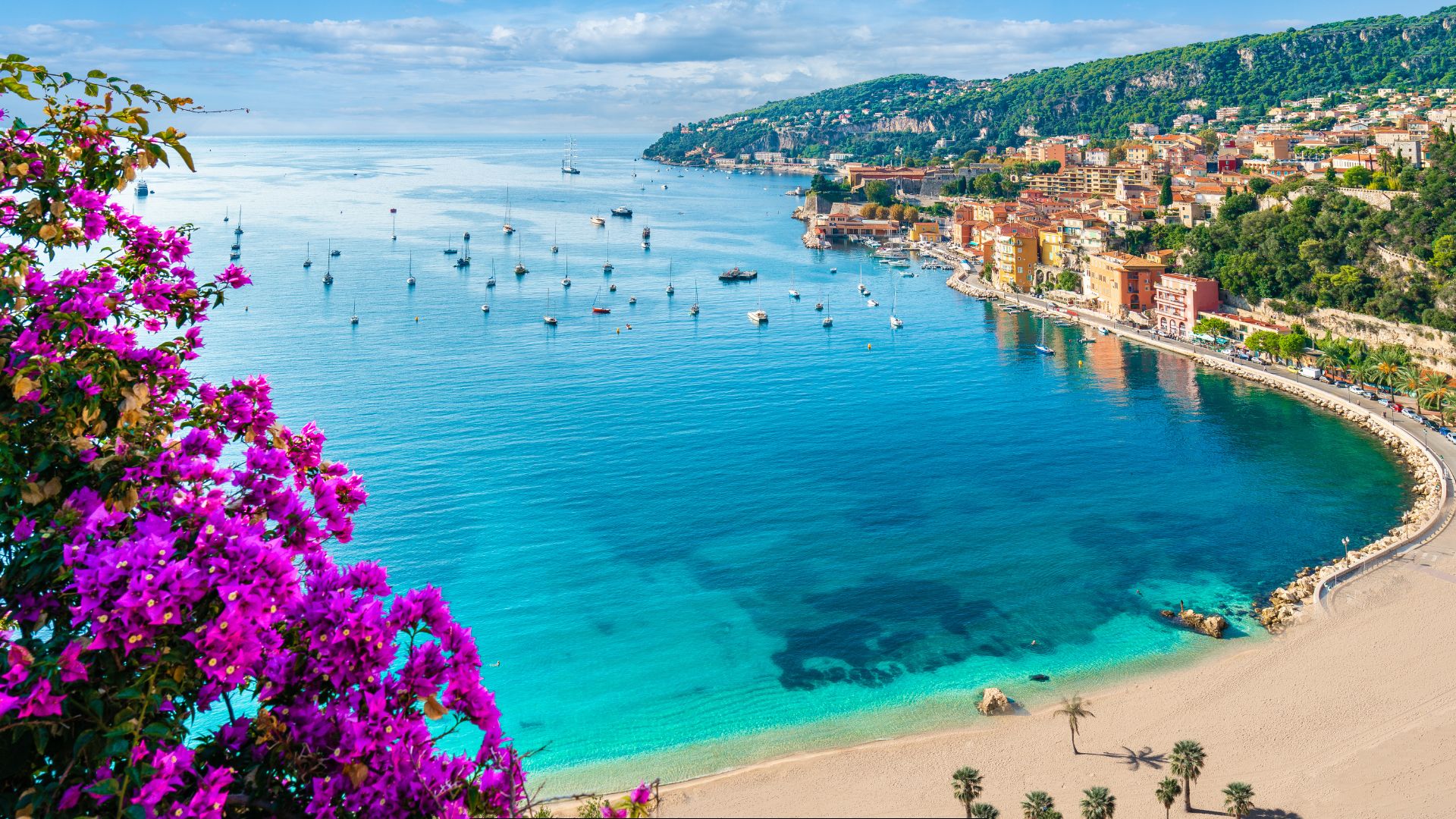 Purple bougainvillea foreground, turquoise bay dotted with sailboats, sandy beach and hillside town