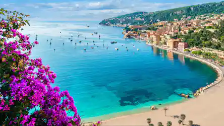 Purple bougainvillea foreground, turquoise bay dotted with sailboats, sandy beach and hillside town