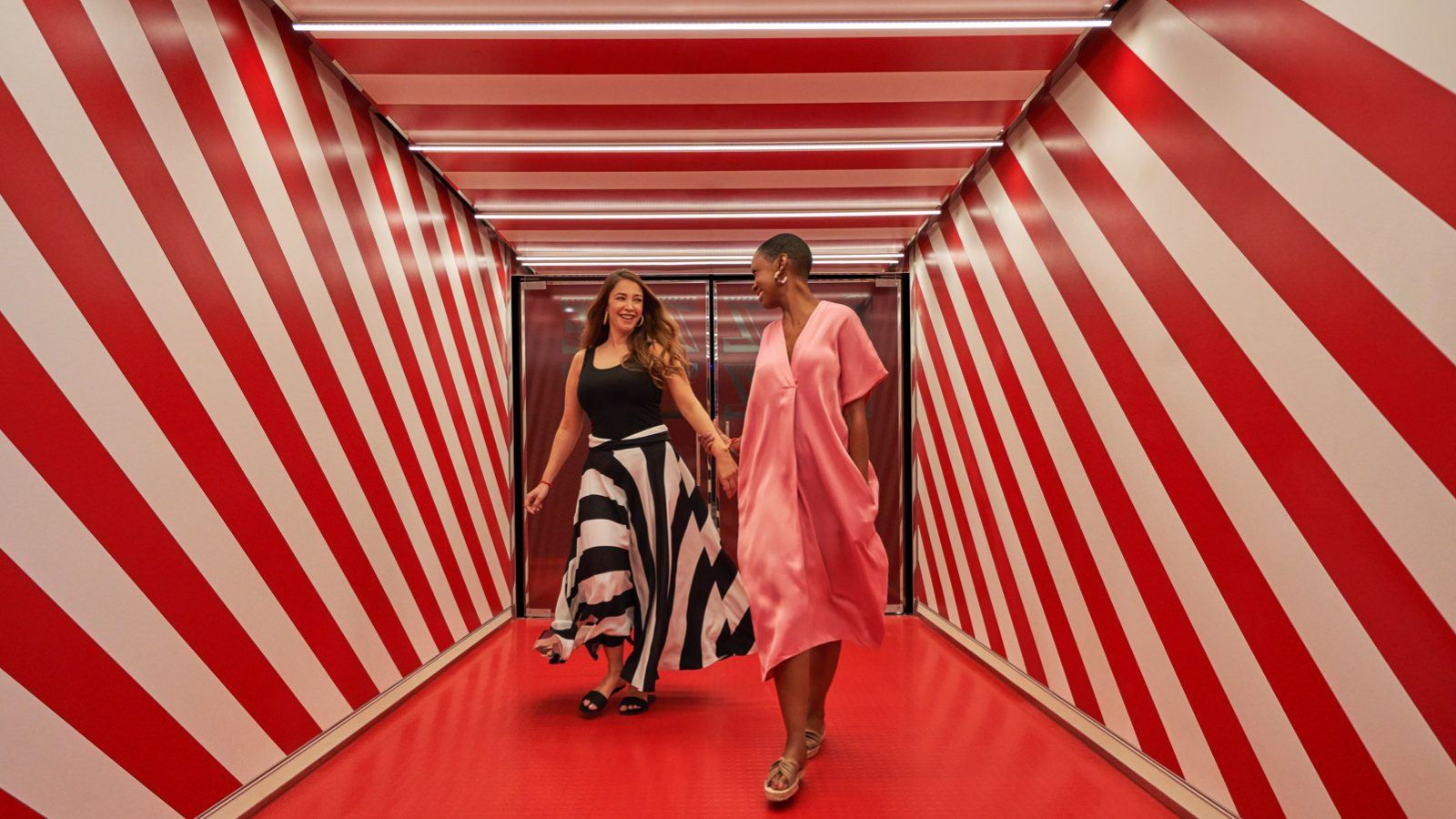 Two smiling women holding hands walking through red-and-white diagonal-striped corridor.