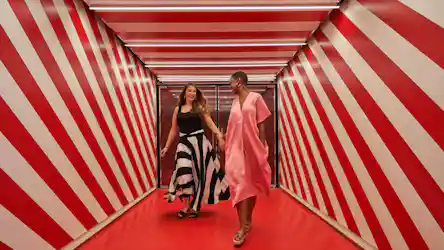 Two smiling women holding hands walking through red-and-white diagonal-striped corridor.