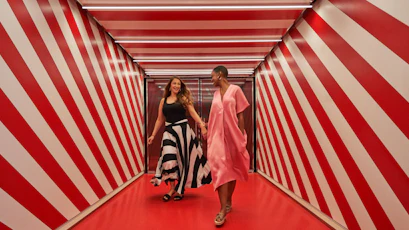 Two smiling women holding hands walking through red-and-white diagonal-striped corridor.
