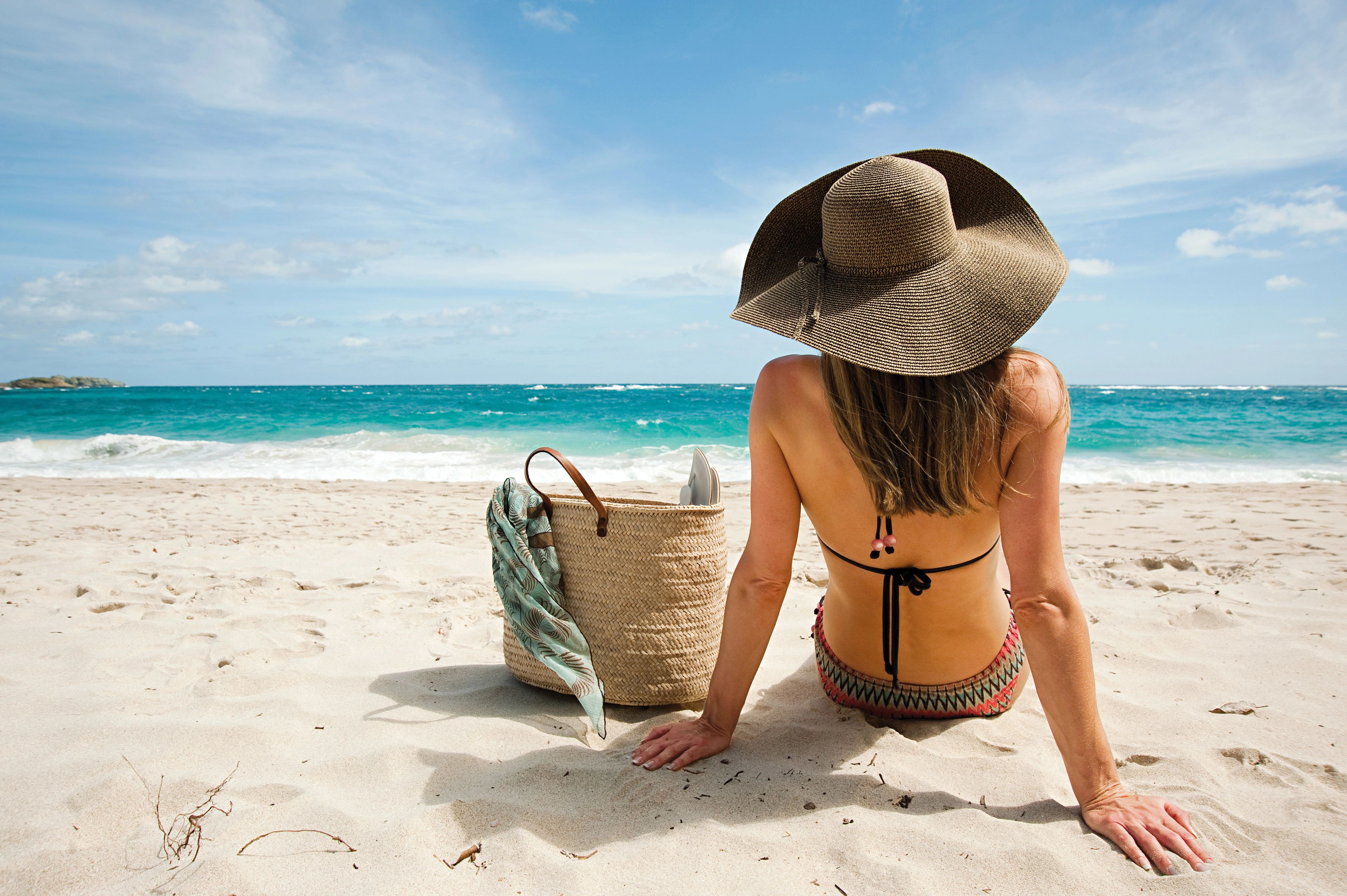 Woman in wide sunhat and bikini sitting on sand beside straw bag, facing turquoise ocean