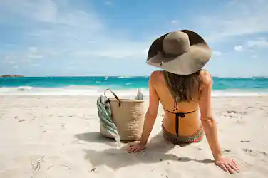 Woman in wide sunhat and bikini sitting on sand beside straw bag, facing turquoise ocean