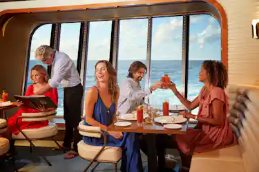 Friends dining on a cruise ship, laughing and toasting beside large ocean windows.