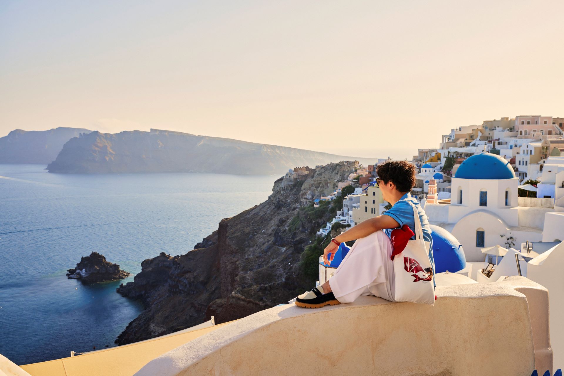 Person seated on Santorini cliff overlooking white houses, blue domes and caldera sea at sunset.