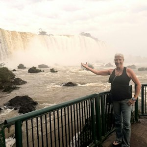 Smiling woman on riverside viewing platform gesturing toward wide misty waterfall.