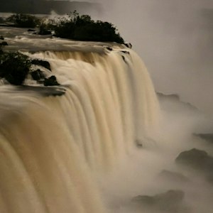 Massive curtain of water plunging over a rocky cliff into mist, vegetation atop the rim