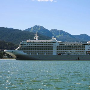 White cruise ship off a forested shore with green mountains and clear blue sky
