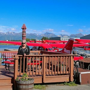 Woman in sunglasses on wooden dock before red floatplanes on a lake with distant snow peaks