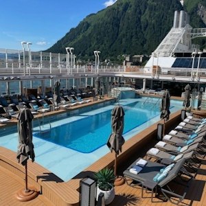 Cruise ship pool deck with rows of lounge chairs, closed umbrellas, and mountain backdrop