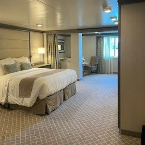 Spacious beige hotel room with king bed, bedside lamp, patterned carpet and seating by window