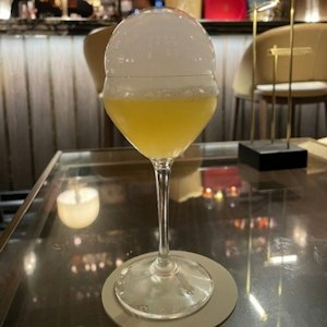 Yellow cocktail in stemmed glass topped by a large white foam bubble, on a glass bar table
