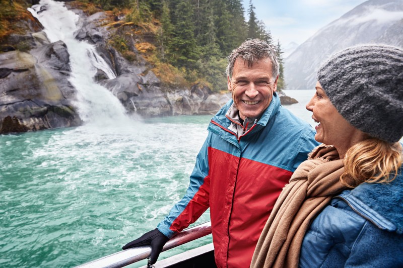 Couple in jackets on boat railing smiling at turquoise water, rocky waterfall and evergreen shore