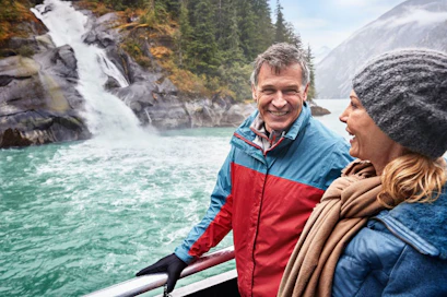 Couple in jackets on boat railing smiling at turquoise water, rocky waterfall and evergreen shore