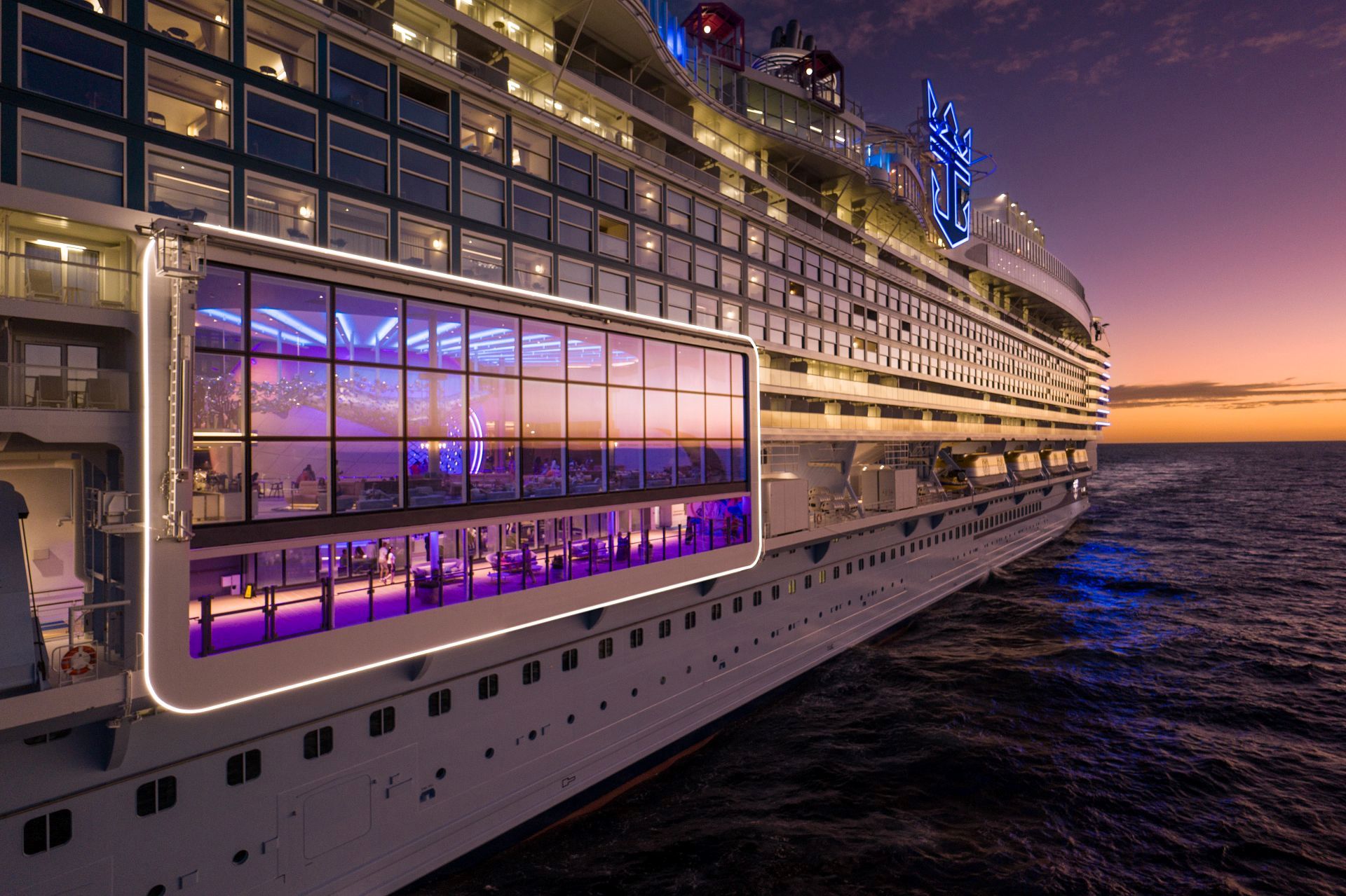 Large cruise ship side with glass-walled lounge and lit balconies at sunset over ocean