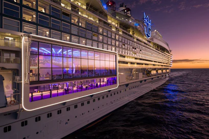 Large cruise ship side with glass-walled lounge and lit balconies at sunset over ocean