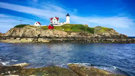 White navigational lighthouse and keeper's house with American flag and red sheds on rocky island