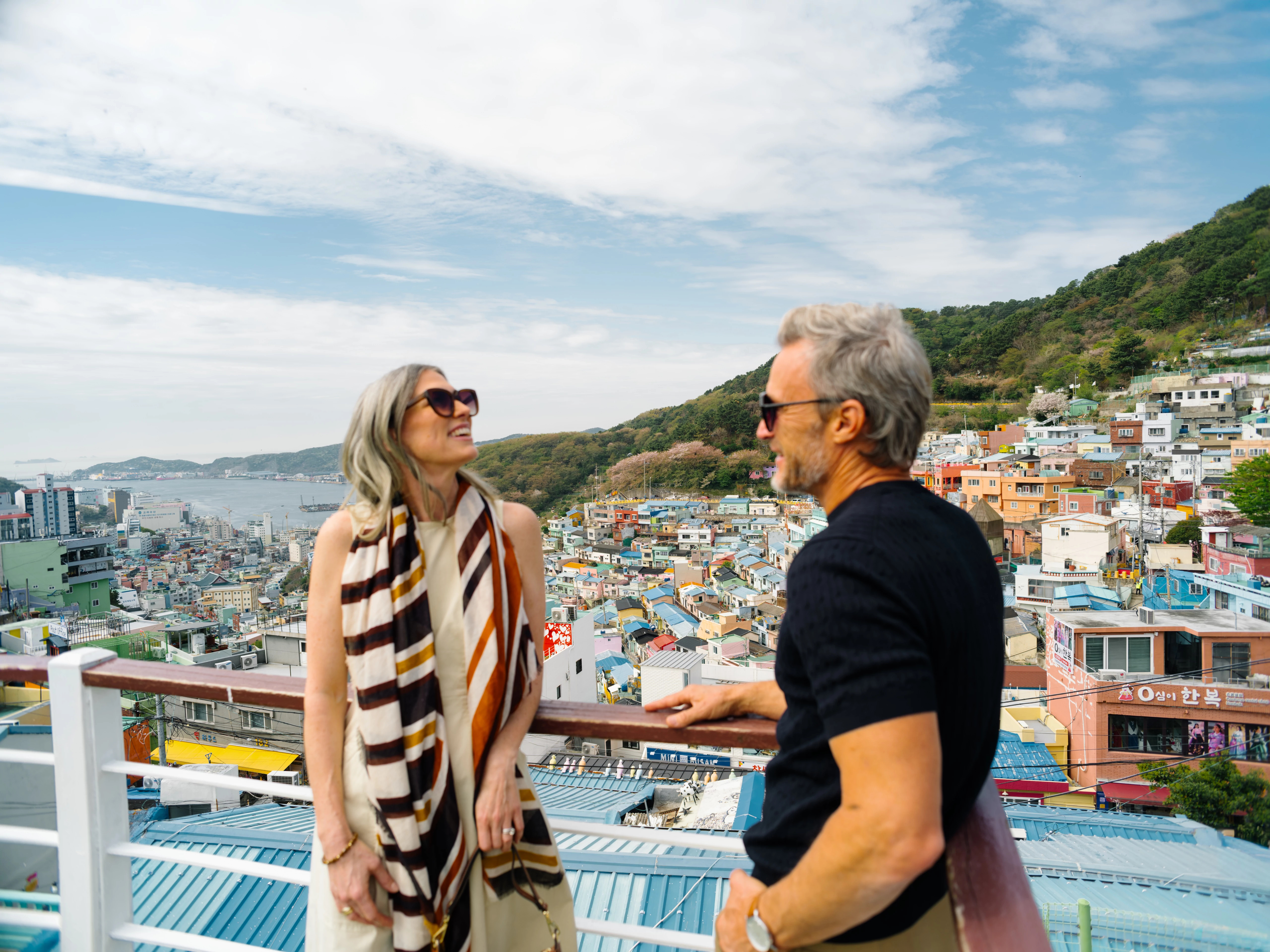 Couple in sunglasses laughing on rooftop overlooking colorful hillside coastal town and harbor