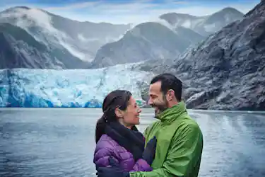 Smiling couple in winter jackets embracing by a glacial lake with blue glacier and mountains