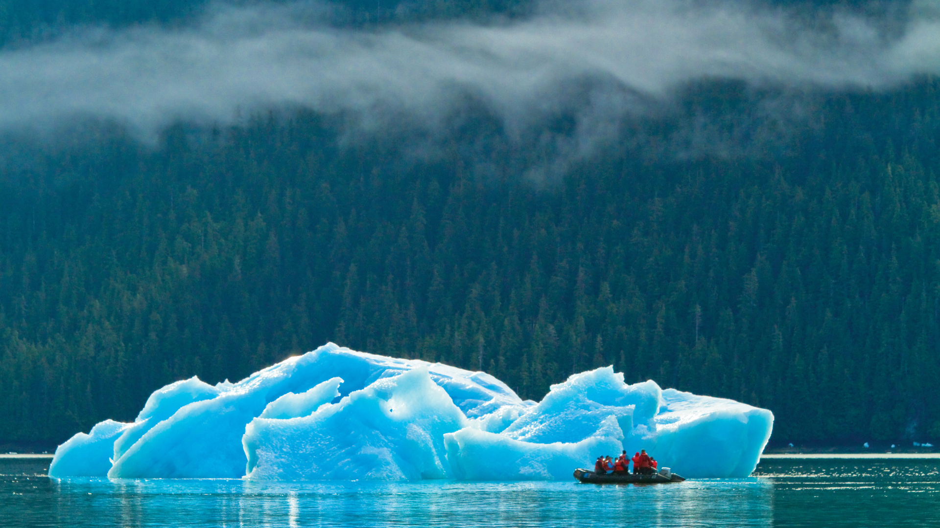 People in a zodiac with red jackets beside a blue iceberg below misty forested mountains