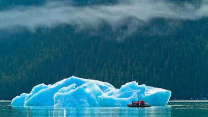 People in a zodiac with red jackets beside a blue iceberg below misty forested mountains