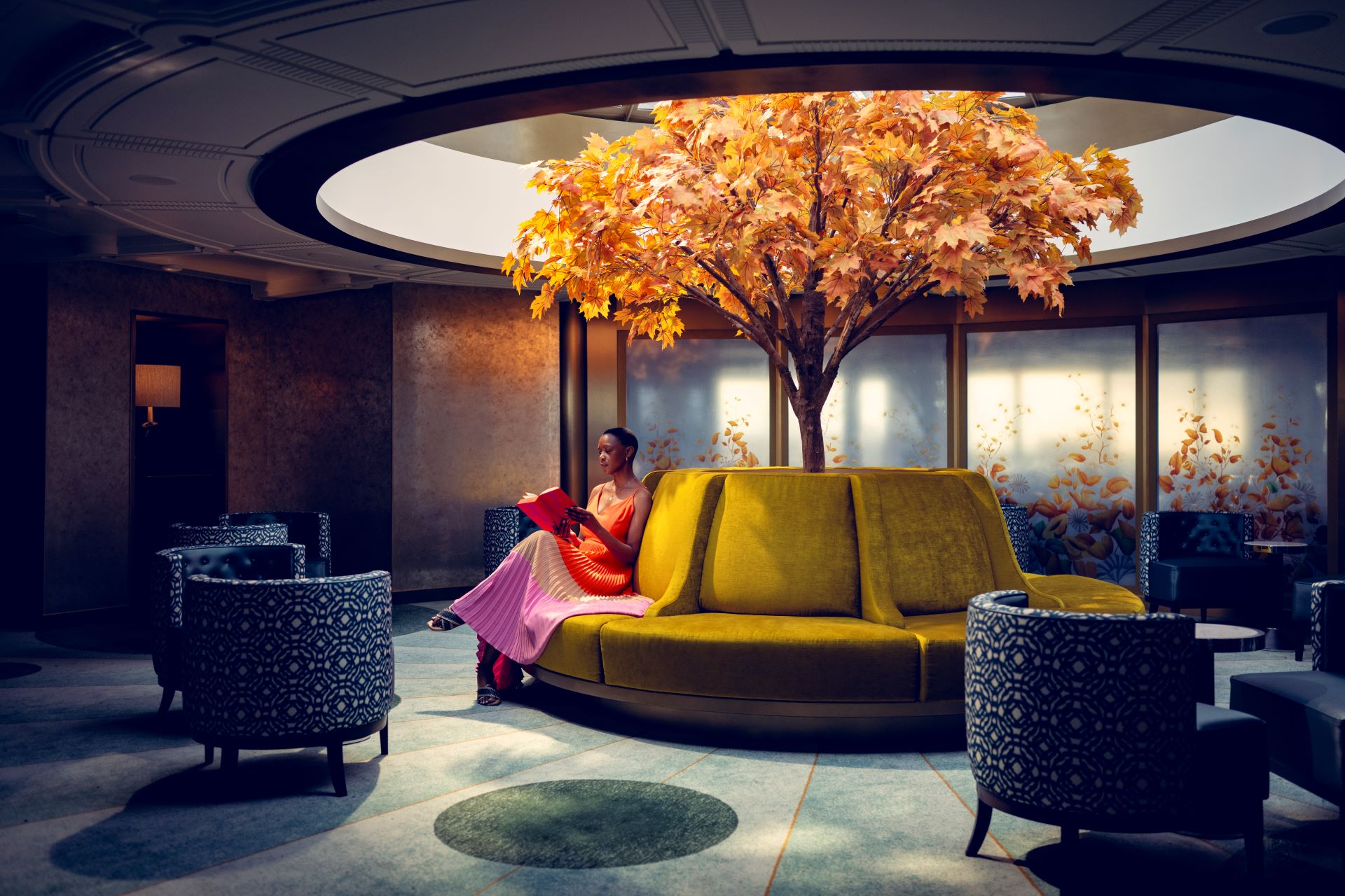 Person reading on circular yellow sofa under indoor orange-leafed tree in a lounge
