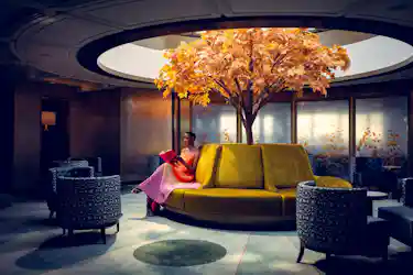 Person reading on circular yellow sofa under indoor orange-leafed tree in a lounge