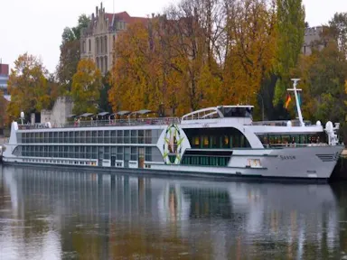 Long white river cruise ship docked beside an autumn tree-lined riverbank and historic building