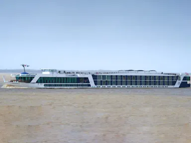 Long white multi-deck river cruise ship with rooftop sun deck and passengers cruising on brown river