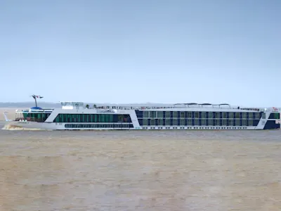 Long white multi-deck river cruise ship with rooftop sun deck and passengers cruising on brown river