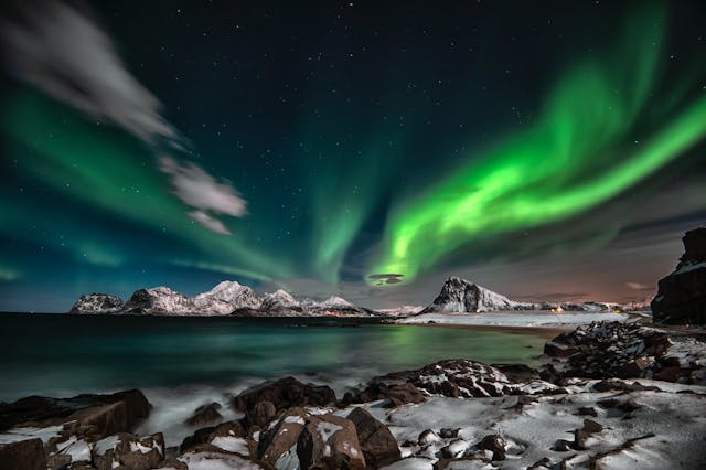 Green northern lights in sky above bay with snowy mountains in distance