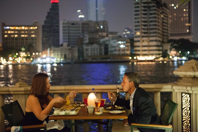 Two people at night at a restaurant eating dinner on a balcony with a view looking over a river to a city skyline.