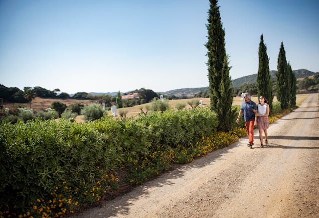 Couple walking arm-in-arm on dirt path next to flowering hedge