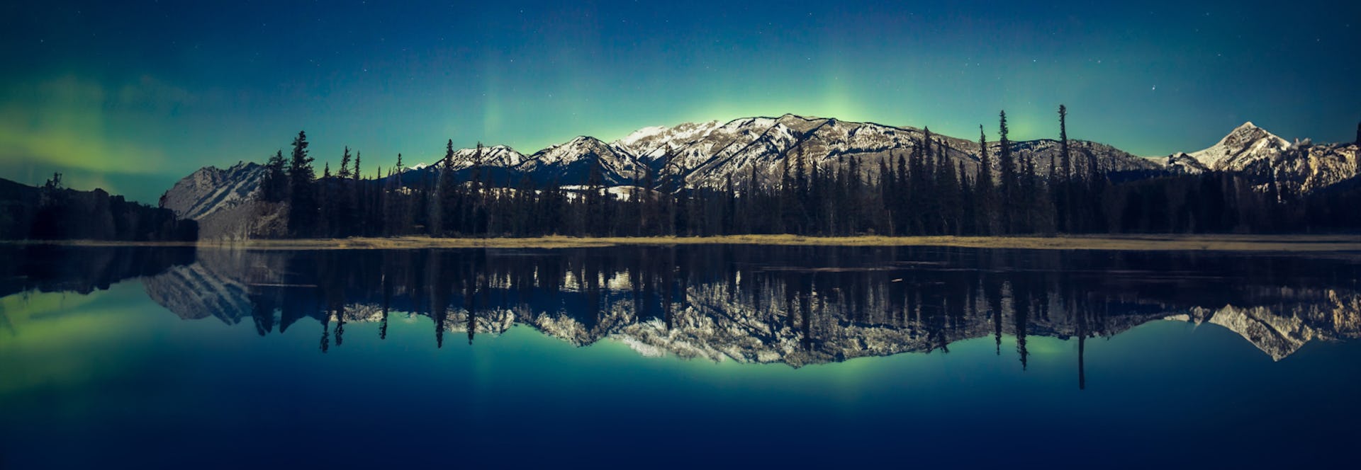 Still lake at night with forest and mountains in background and Aurora borealis shining in sky